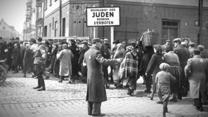 Židovský policista a německý voják usměrňují provoz na hlavní ulici, která rozdělovala ghetto. Později byl na této křižovatce postaven dřevěný most. U vchodu do ghetta je v němčině Židovská obytná oblast, vstup zakázán. Foto USHMM/Wikimedia Commons