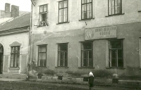 Budova bývalé tiskárny s nápisem: "LIDOVÉ DRUŽSTVO ODĚVNÍ",  v okně Anna Krčmařová s manželem Eduardem Krčmařem nejstařším, dole vnučka Dagmar, asi 1957