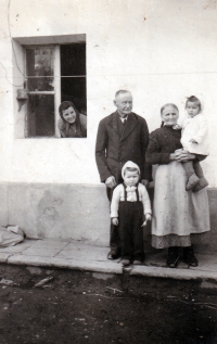 Monika Ruská with her grandfather František Theuer, her younger sister, their neighbor and her mother (in the window) on their family farm in Bolatice / around 1949